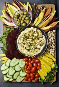 large platter of beets, cucumber slices, cherry tomatoes, endive spreads, rainbow baby carrots, and olives surrounding bowl of pistachio yogurt dip