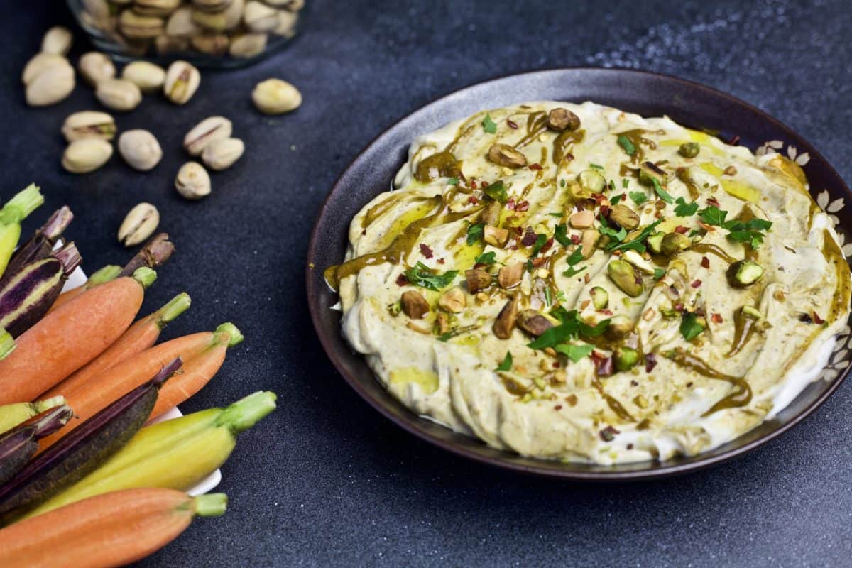 bowl of pistachio yogurt dip next to platter of rainbow baby carrots and in-shell pistachios in background