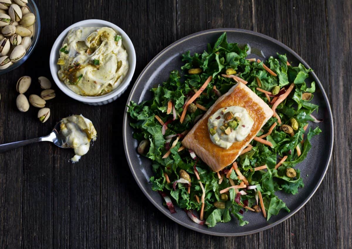 plate of mixed greens with roasted salmon featuring dollop of pistachio yogurt dip on top and bowl of sauce in corner