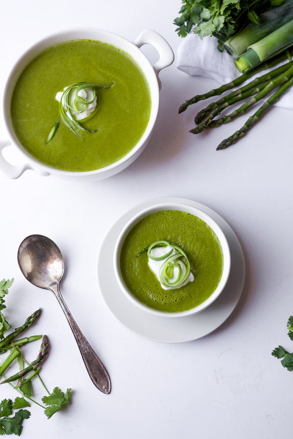 spring leek and asparagus soup in small bowl and with seasonal ingredients like asparagus leeks and herbs surrounding